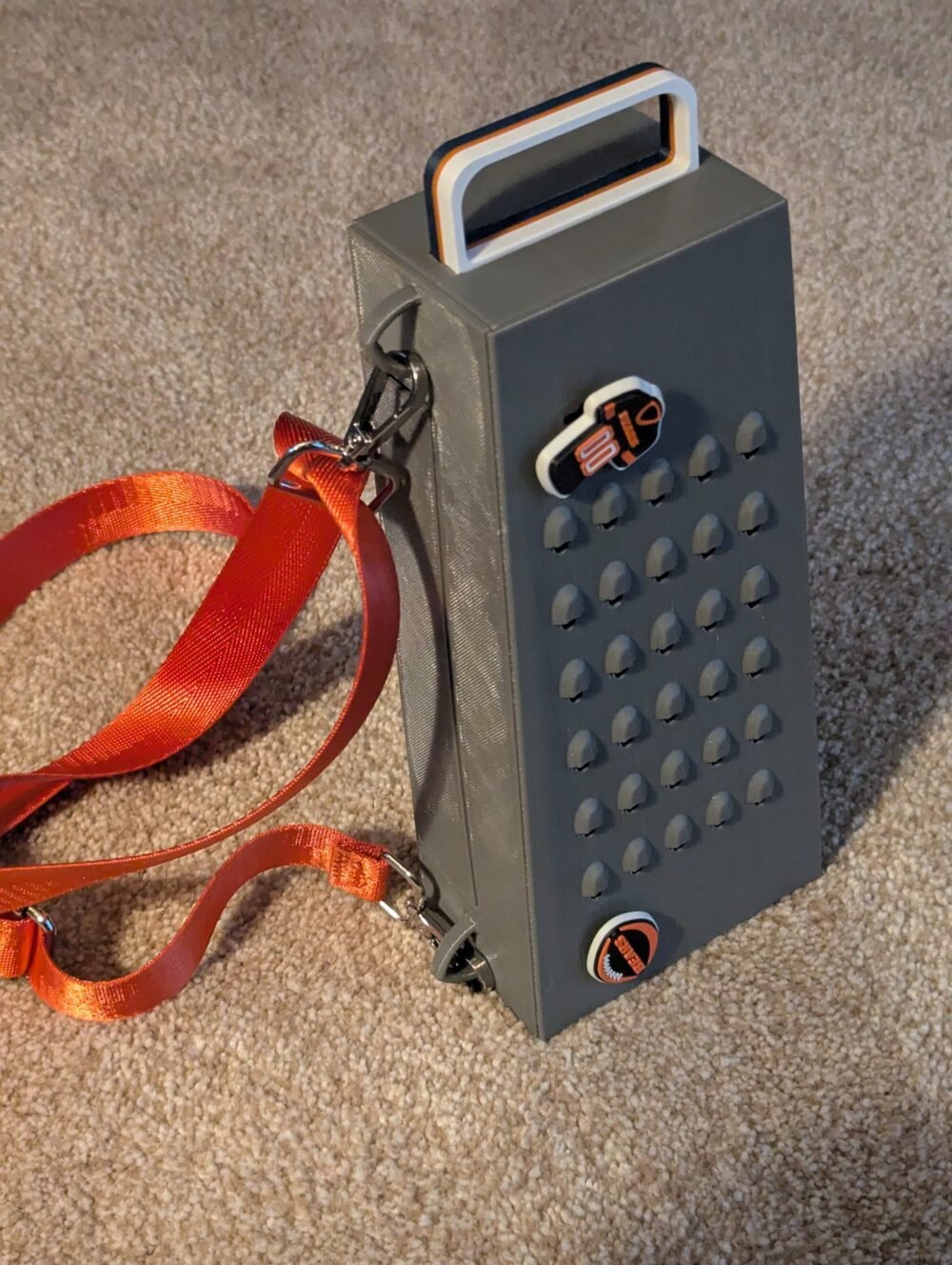 The Grater Purse — a gray cheese-grater-shaped handbag with a top handle and orange crossbody strap