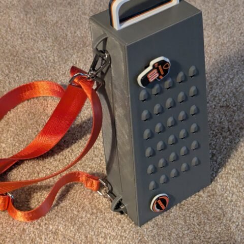 The Grater Purse — a gray cheese-grater-shaped handbag with a top handle and orange crossbody strap